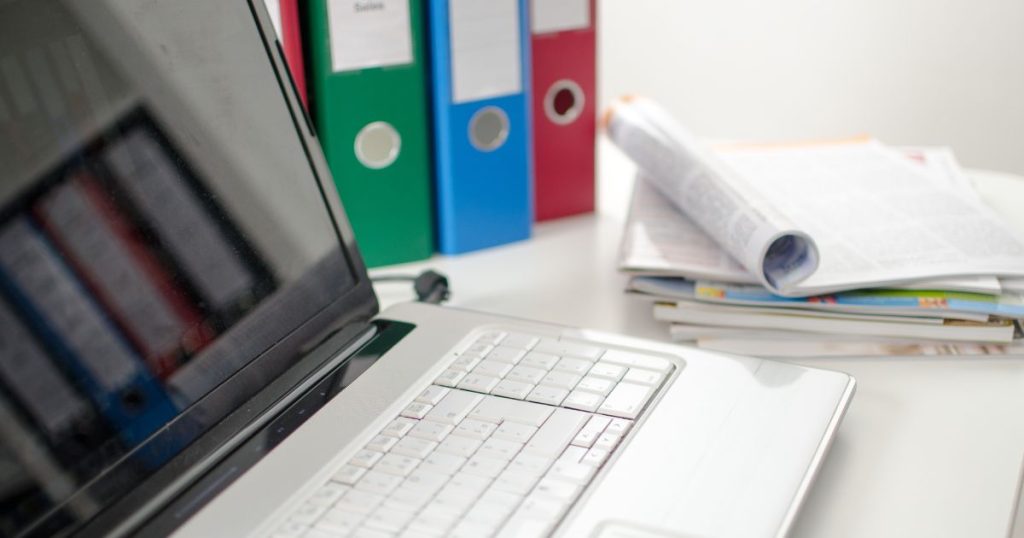 Computer and binders with a stack of papers on a desk.